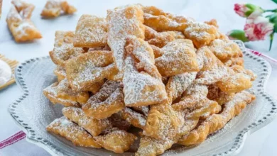 Prato decorado com enroladinho de banana fritos, dourados e crocantes, polvilhados com açúcar de confeiteiro, sobre uma toalha listrada vermelha com flores ao fundo