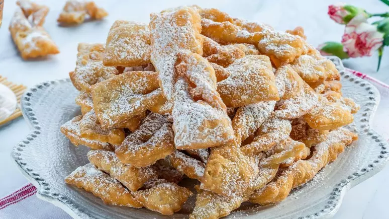 Prato decorado com enroladinho de banana fritos, dourados e crocantes, polvilhados com açúcar de confeiteiro, sobre uma toalha listrada vermelha com flores ao fundo