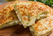 Pedaço de Focaccia de Cebola Caramelizada e Queijo, com crosta dourada, servida sobre uma tábua de madeira com ingredientes verdes ao fundo.