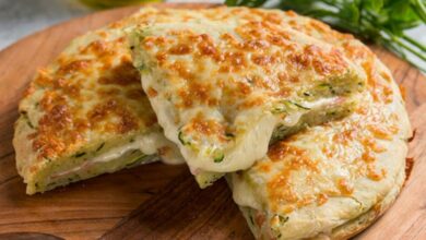 Pedaço de Focaccia de Cebola Caramelizada e Queijo, com crosta dourada, servida sobre uma tábua de madeira com ingredientes verdes ao fundo.