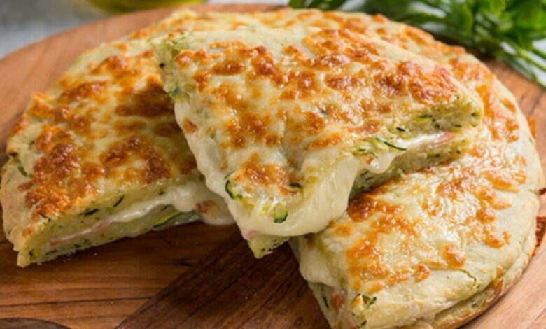 Pedaço de Focaccia de Cebola Caramelizada e Queijo, com crosta dourada, servida sobre uma tábua de madeira com ingredientes verdes ao fundo.