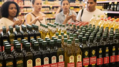 Uma prateleira de supermercado cheia de garrafas de azeite e outros óleos, algumas delas com etiquetas vermelhas indicando "PRODUTO SUSPENSO". Ao fundo, embaçados, quatro consumidores parecem discutir ou observar os produtos, com o foco principal nas garrafas de azeite e nas etiquetas de suspensão.