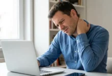 Foto de um homem com expressão de dor segurando seu pescoço travado, sentado em uma mesa de escritório com um notebook e um celular à sua frente, ilustrando o desconforto causado pelo uso excessivo de telas.
