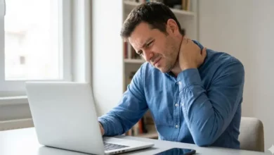 Foto de um homem com expressão de dor segurando seu pescoço travado, sentado em uma mesa de escritório com um notebook e um celular à sua frente, ilustrando o desconforto causado pelo uso excessivo de telas.