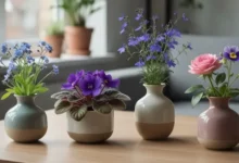 Flores pequenas para decorar - Fotografia realista em alta qualidade de uma mesa de madeira clara próxima a uma janela iluminada, com cinco pequenos vasos de cerâmica contendo as seguintes flores: miosótis azul com centro amarelo, violeta-africana roxa de folhas aveludadas, lobélia azul-violeta delicada, mini-rosa em tom rosado e torênia com pétalas roxas e amarelas.