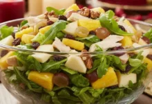 Uma vibrante Salada Tropical de Natal servida em uma tigela de vidro transparente sobre uma mesa de ceia decorada. A salada contém alface americana, rúcula, cubos de abacaxi, maçã, uvas passas, nozes e frango desfiado com molho cremoso, destacando suas cores frescas e suculentas. O fundo mostra velas acesas e decorações natalinas em vermelho e dourado, criando um ambiente elegante e festivo.