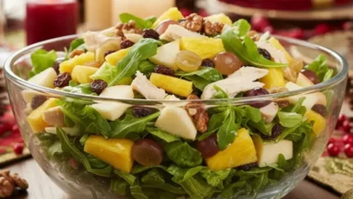 Uma vibrante Salada Tropical de Natal servida em uma tigela de vidro transparente sobre uma mesa de ceia decorada. A salada contém alface americana, rúcula, cubos de abacaxi, maçã, uvas passas, nozes e frango desfiado com molho cremoso, destacando suas cores frescas e suculentas. O fundo mostra velas acesas e decorações natalinas em vermelho e dourado, criando um ambiente elegante e festivo.