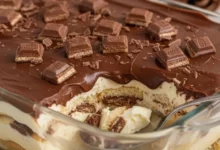 Uma fotografia de alta qualidade de um deliciosa receita de pavê de Bis servido em um refratário de vidro elegante. O pavê deve mostrar camadas bem definidas de creme suave, biscoitos e pedaços generosos de chocolate Bis. A cobertura deve ser uma ganache de chocolate brilhante, com alguns pedaços de Bis inteiros ou triturados por cima para decorar. Foco em close-up, com uma colher inserida suavemente no pavê, sugerindo a textura cremosa. O fundo deve ser levemente desfocado, com um toque de decoração natalina sutil (talvez um pisca-pisca suave ou ramos verdes) para indicar a temática de fim de ano. Iluminação quente e convidativa, cores ricas e contrastantes.