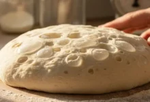 Massa de pizza caseira com grandes bolhas de ar sobre bancada de madeira, mostrando a fermentação lenta. A luz do sol incide sobre a massa, realçando a textura leve.