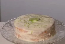 Bolo de limão fofinho com cobertura cremosa branca de limão e raspas verdes, servido em uma mesa de café da tarde iluminada.
