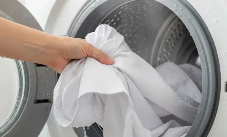 Mão retirando uma camisa branca impecável de dentro da máquina de lavar após clarear roupas brancas.