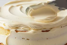 Espátula espalhando uma camada generosa de glace de leite condensado sobre um bolo alto, destacando a textura lisa e cremosa.