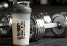 Shakeira com Whey Protein para Hipertrofia Muscular ao lado de pesos de academia, destacando a suplementação para ganho de massa.